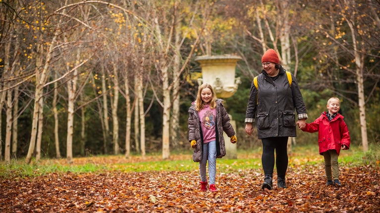 A woman and two children, dressed in warm coats and bobble hats, walking through fallen leaves with autumn trees and a garden statue in the background